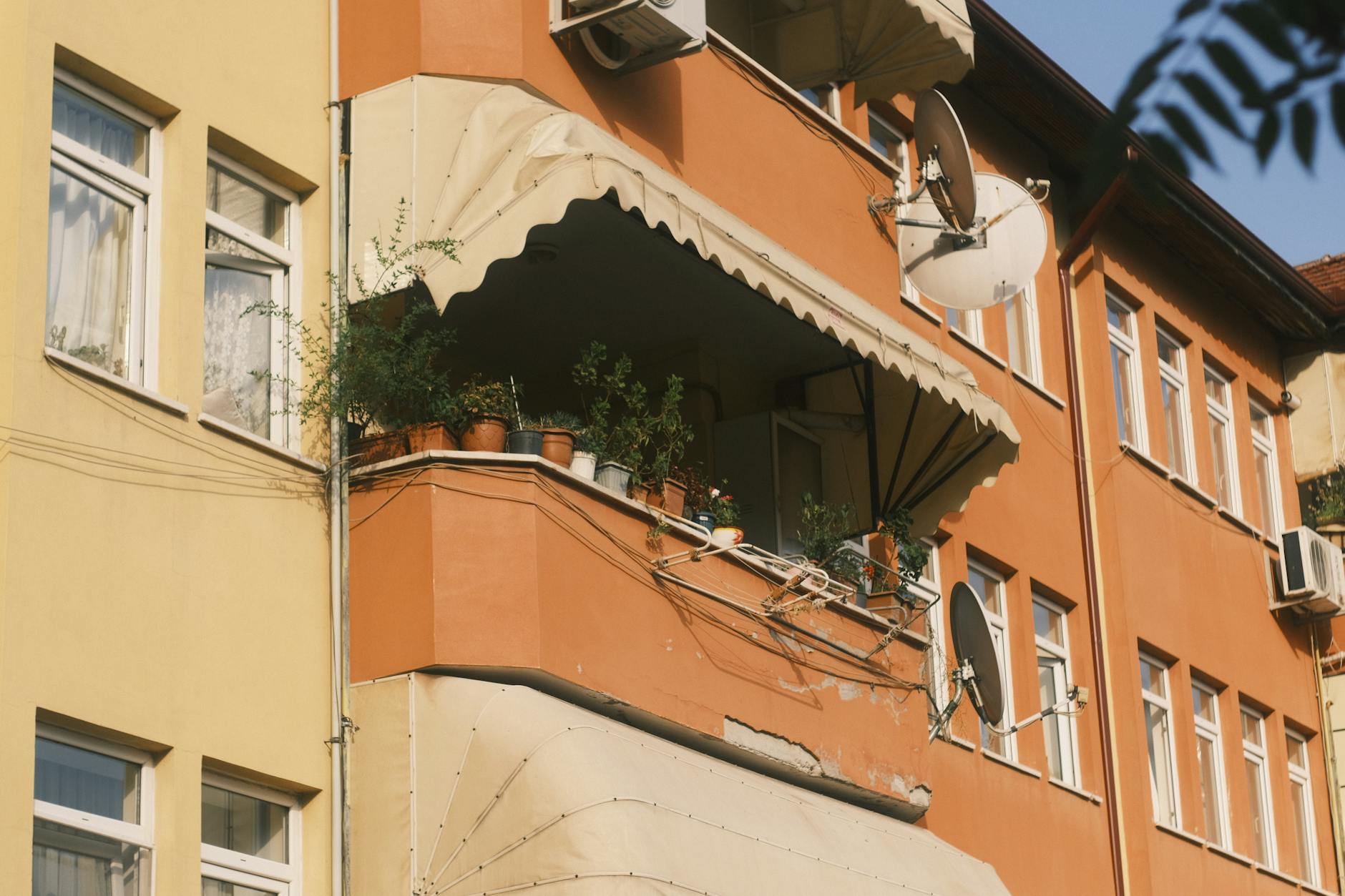 Exterior view of an orange building with potted plants on the balcony and satellite dishes.