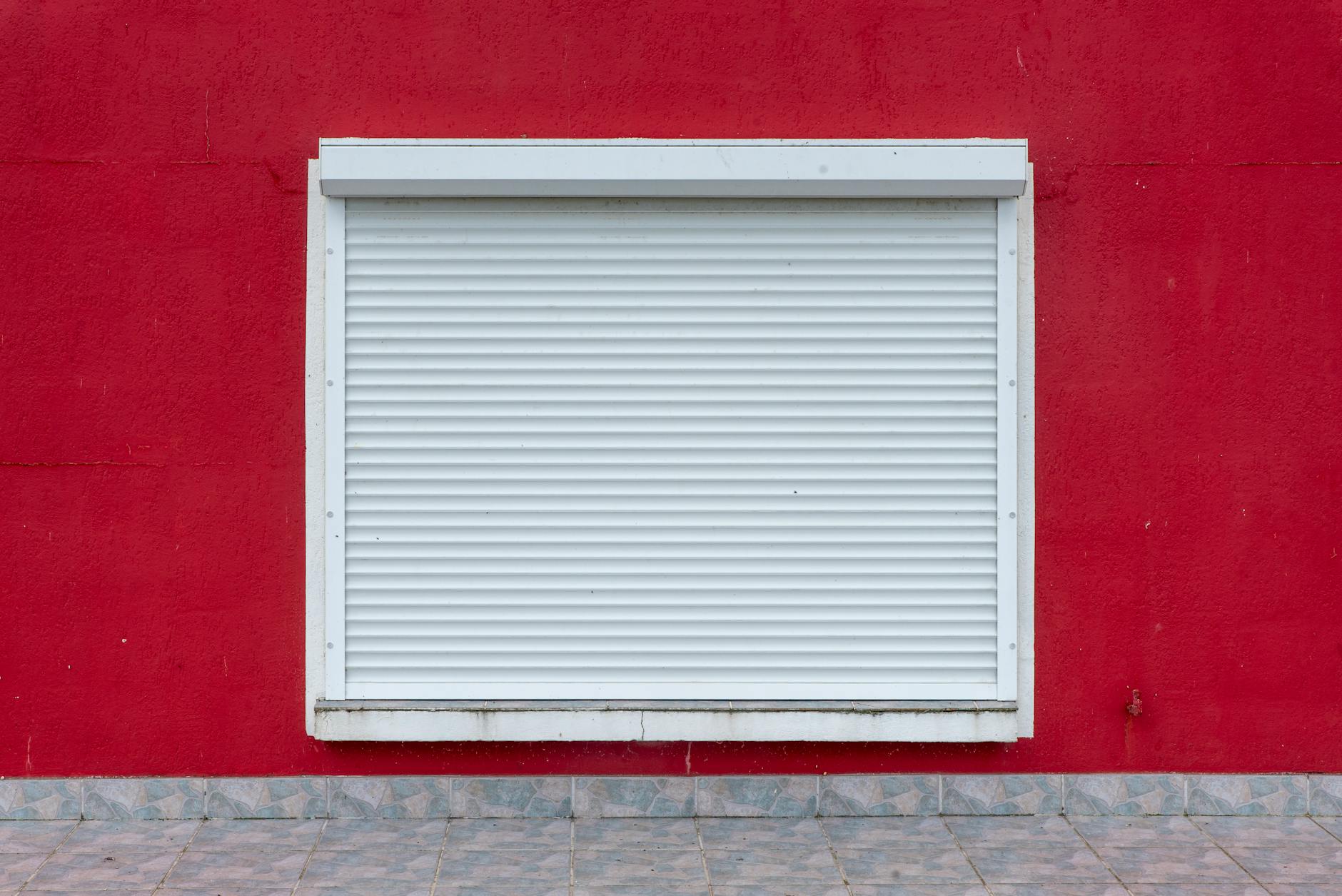 A minimalist exterior view of a white roller shutter on a vibrant red wall.